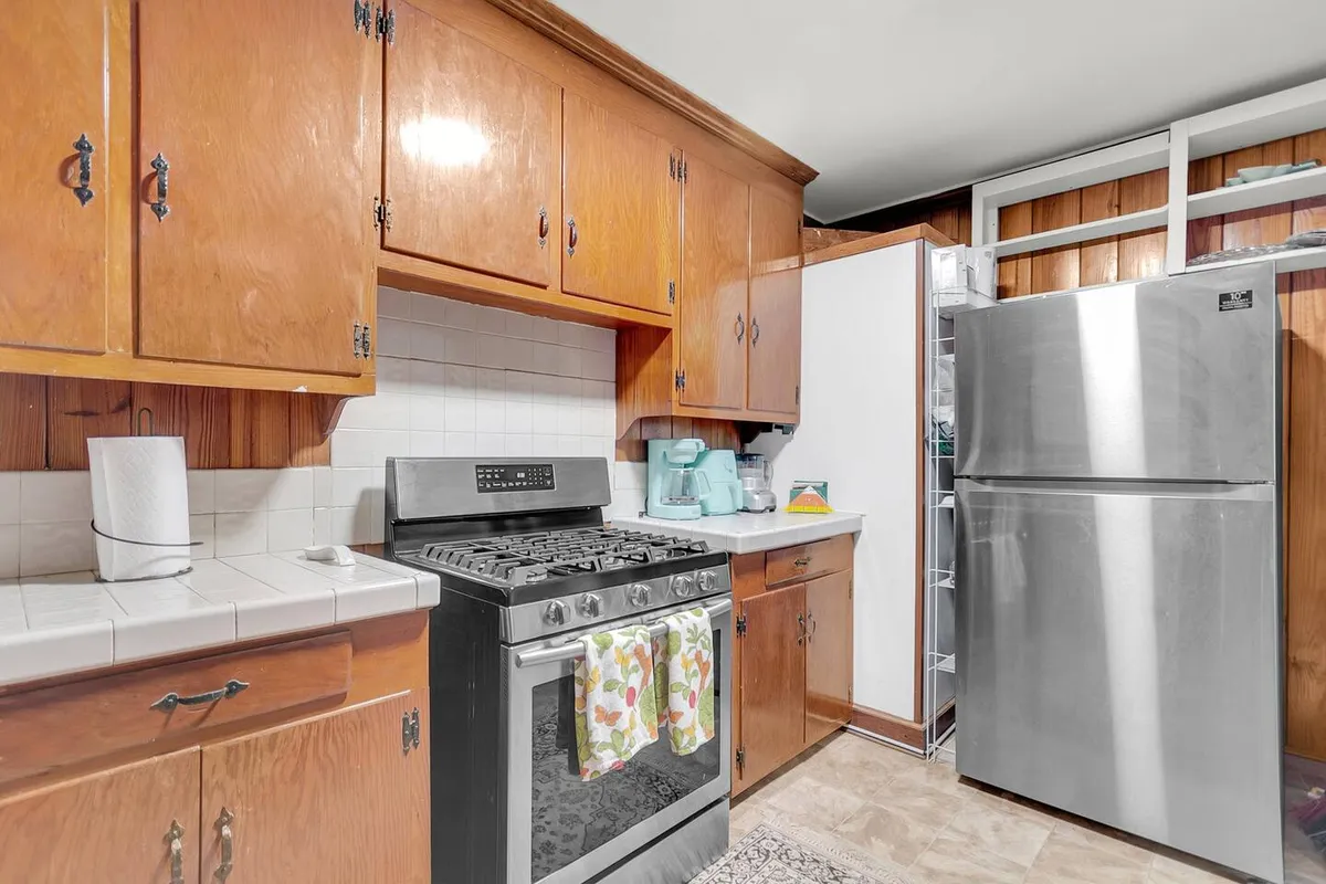 A warm, homely kitchen featuring a stainless steel refrigerator, gas stove, wooden cabinets, and a colorful dish towel hanging by the oven. The kitchen is well-lit and inviting, with plenty of space for cooking and meal preparation, making it ideal for family gatherings and culinary activities.