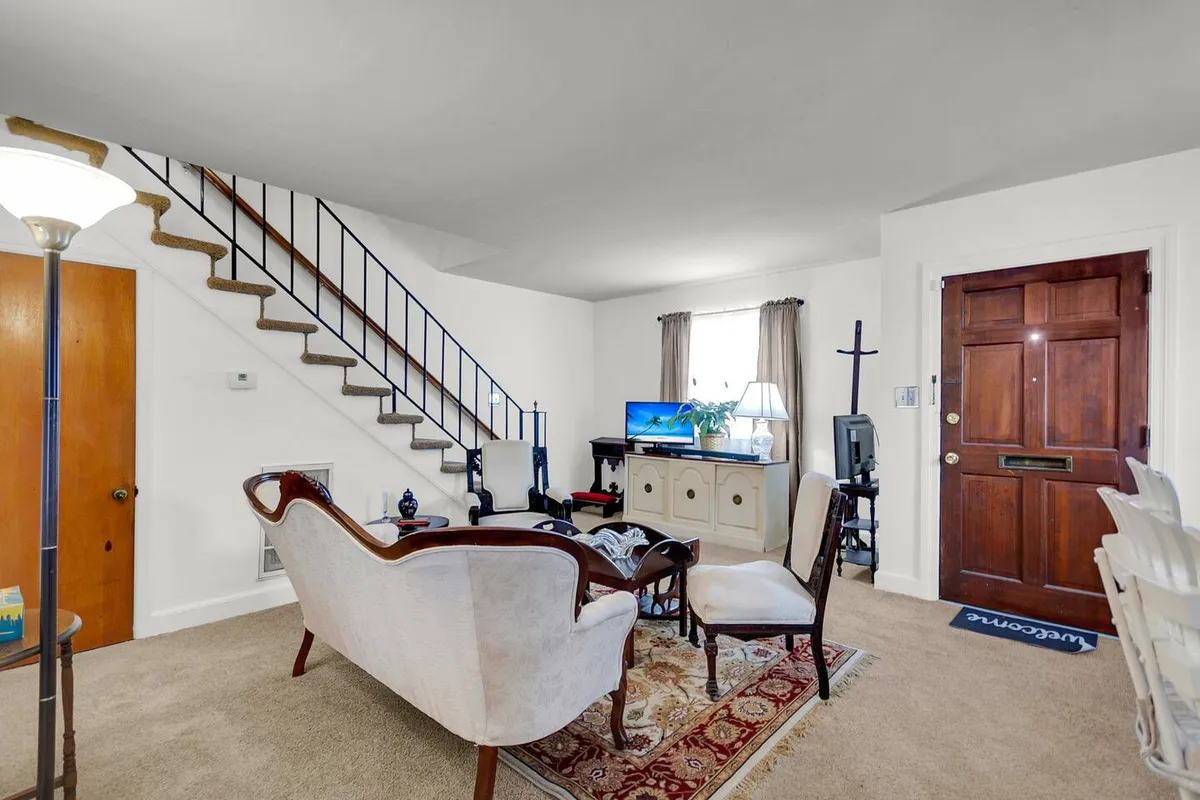 A comfortable living room featuring a plush sofa, stairs leading to the upper floor, a TV, and a visible entrance door. The space is filled with natural light from large windows, creating a bright and inviting atmosphere suited for socializing, movie nights, or casual relaxation.