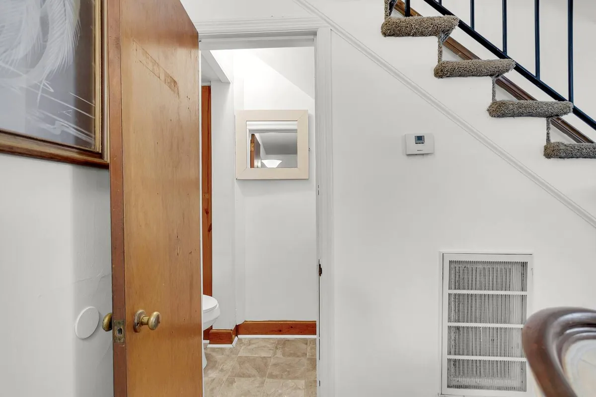 An image of the home’s interior hallway showing a wooden door, staircase, and a visible bathroom entrance with a mirror. This perspective highlights the bathroom’s accessibility and its convenient location for everyday use.