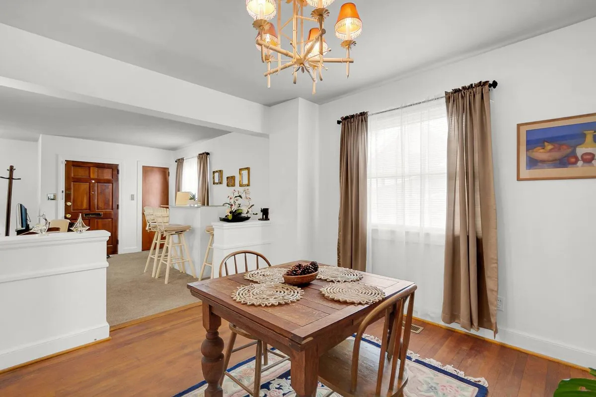 A bright and welcoming dining area centered around a wooden dining table, illuminated by a chandelier and natural light streaming through a window. Decorative wall art and comfortable seating make this a perfect spot for family meals and lively conversations.