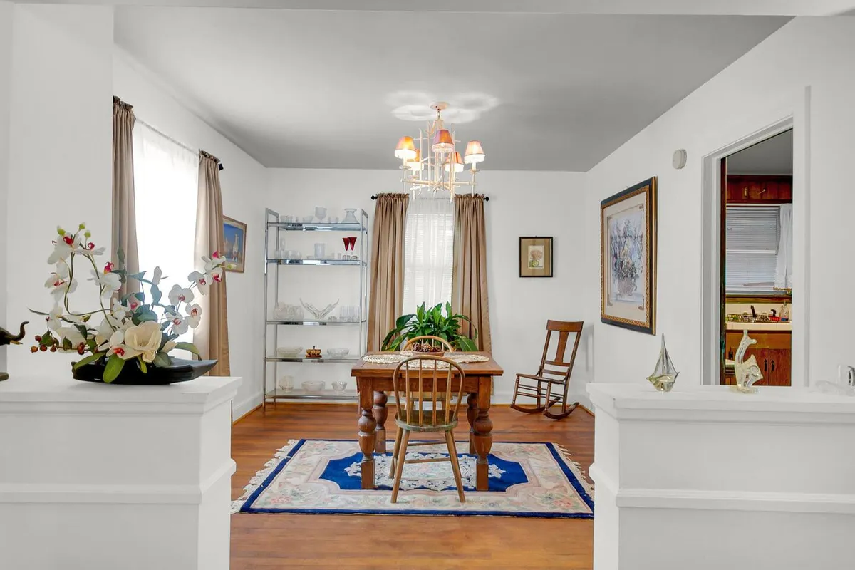 A welcoming dining area is anchored by a central table and illuminated by an elegant chandelier. The space is cozy and versatile, with a display shelf and a rocking chair offering a relaxed setting for meals, remote work, or casual meetings. Sunlight brightens the room, highlighting its inviting atmosphere.