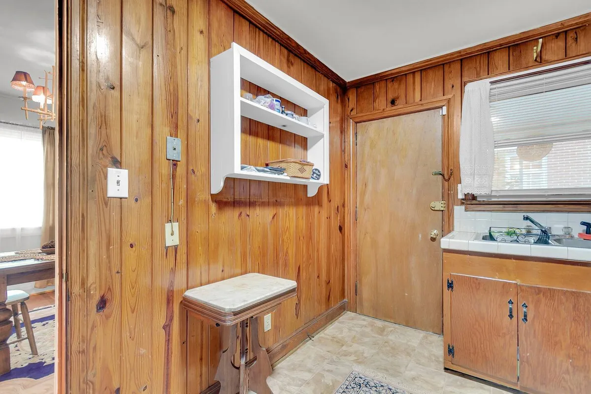 This image features a kitchen area with wood-paneled walls, a white shelving unit, a door leading outside, and a kitchen counter with a sink. The space is bathed in natural light, highlighting areas for preparing and sharing meals.