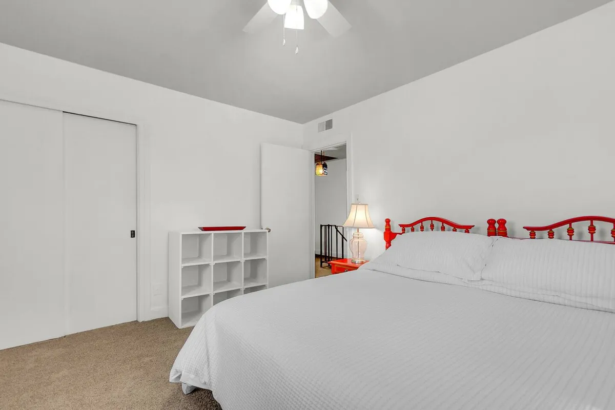 The image presents a calm and cozy bedroom with a red bed frame, white bedding, a nightstand with a lamp, and a shelving unit. The setup suggests a perfect spot for unwinding with a book or enjoying a morning coffee in bed.