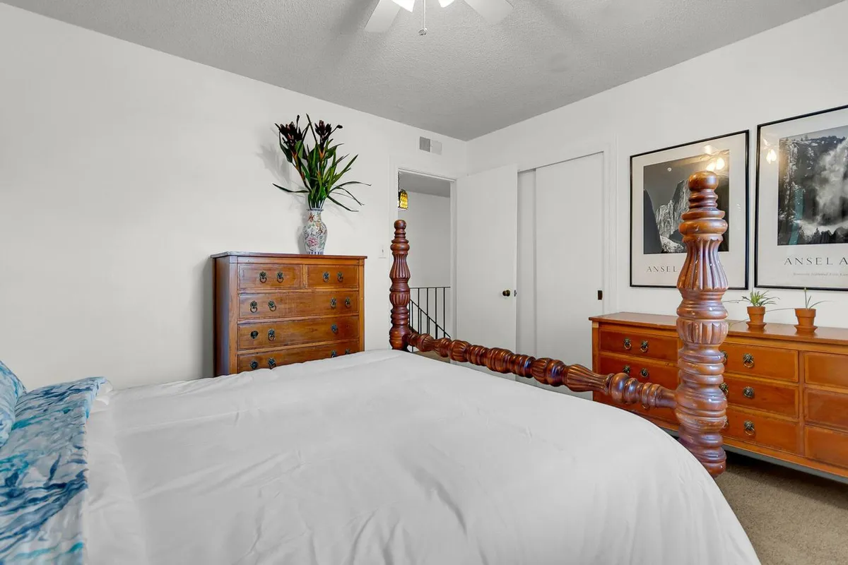 This image showcases a tranquil bedroom with a wooden bed frame, a chest of drawers, a decorative vase with flowers, and artwork adorning the wall. Soft natural light fills the space, highlighting the peaceful atmosphere and thoughtful decor.