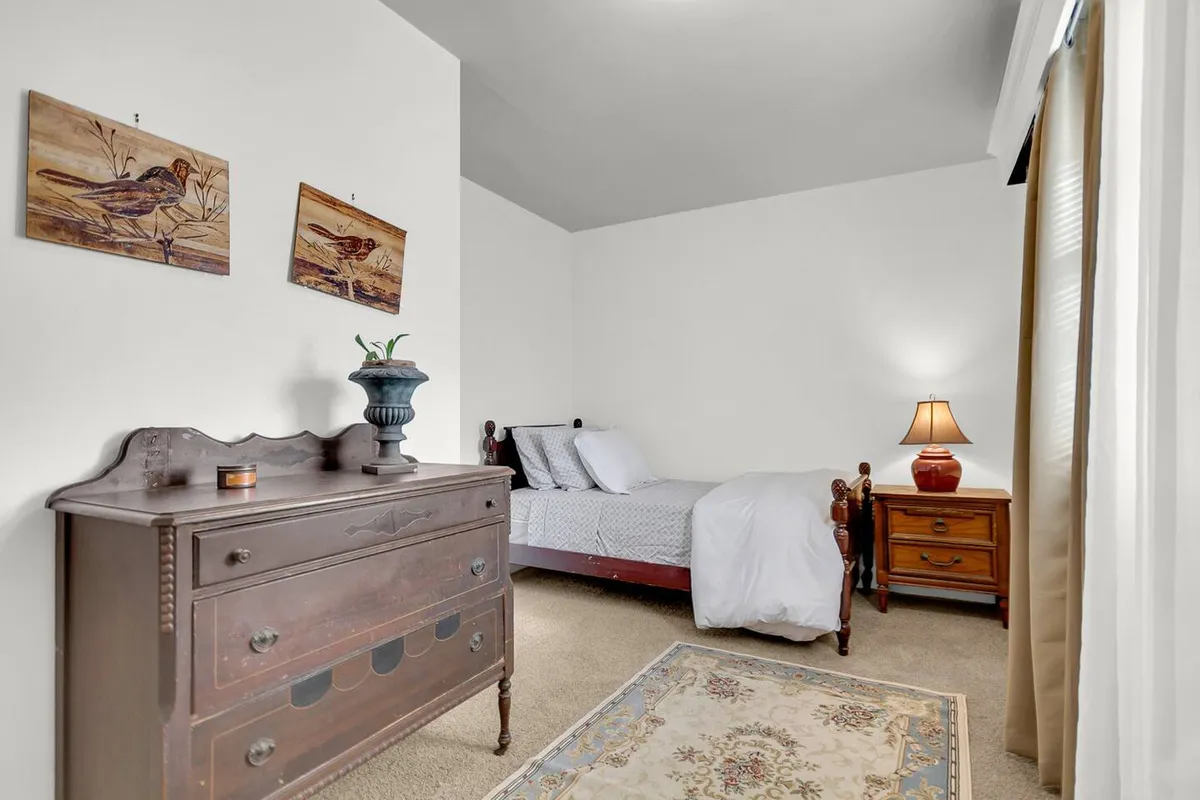 A peaceful bedroom is depicted, featuring a comfortable bed, a dresser, a lamp, and curated artwork. Morning sunlight streams through the window, creating a serene and inviting place for rest and relaxation.